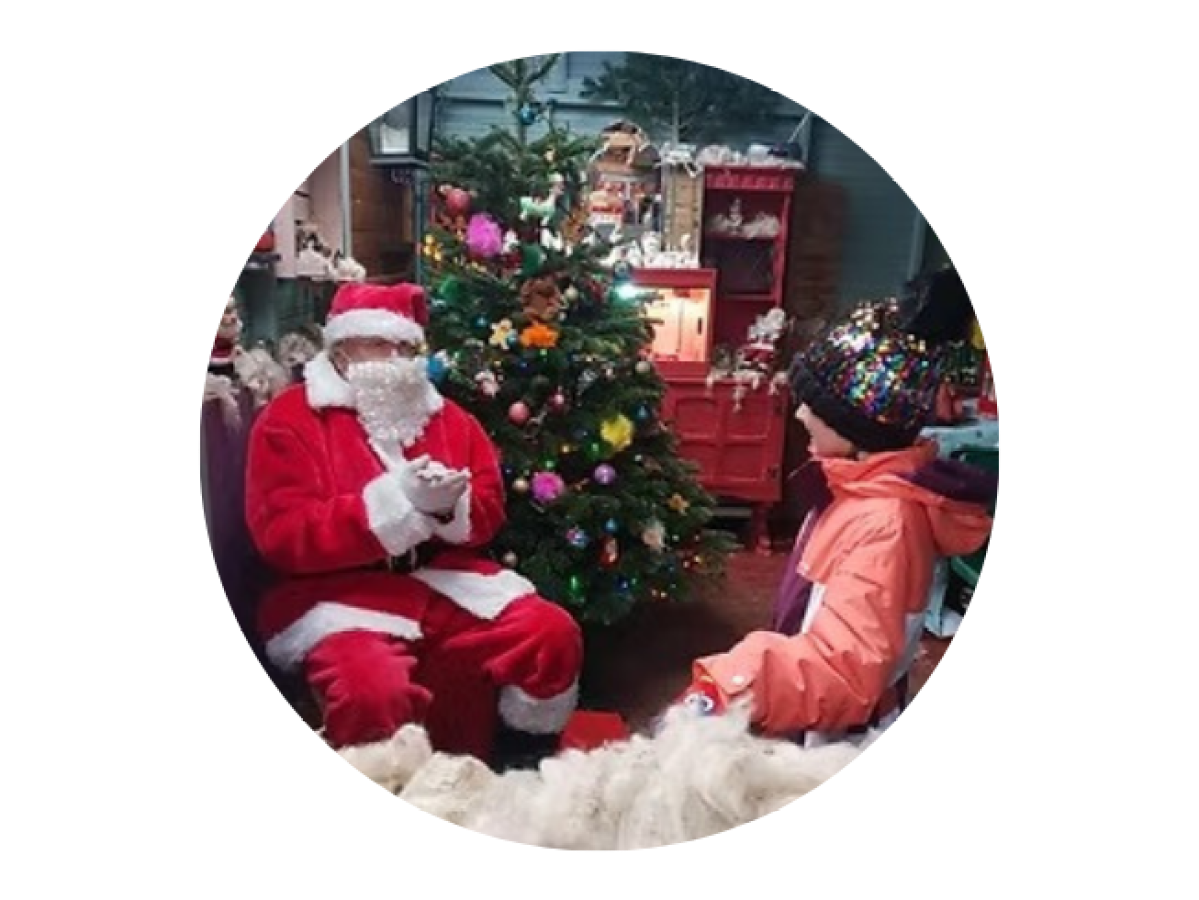Santa Claus sitting by a decorated Christmas tree, talking to a child in winter clothes.