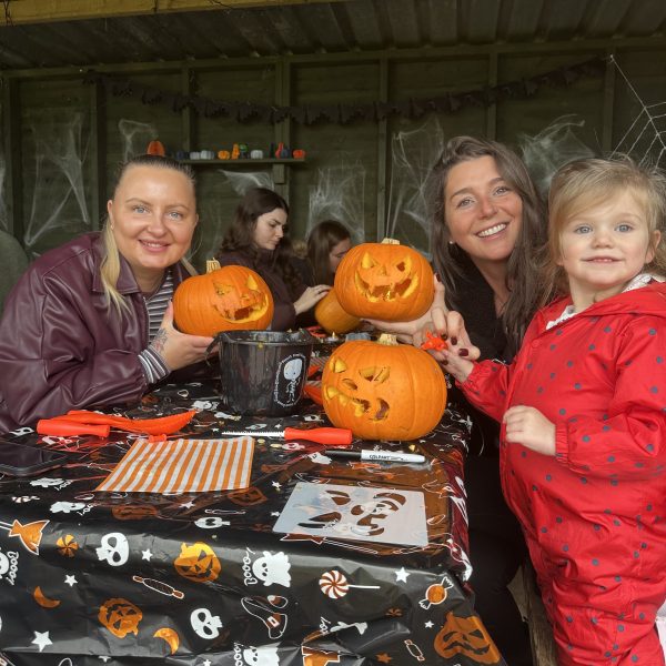 Three people carving pumpkins with a Halloween-themed tablecloth in a decorated room.