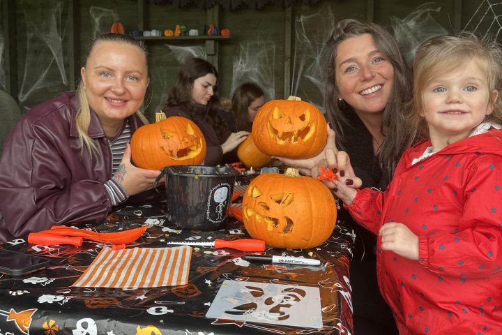 Three people carving pumpkins with a Halloween-themed tablecloth in a decorated room.