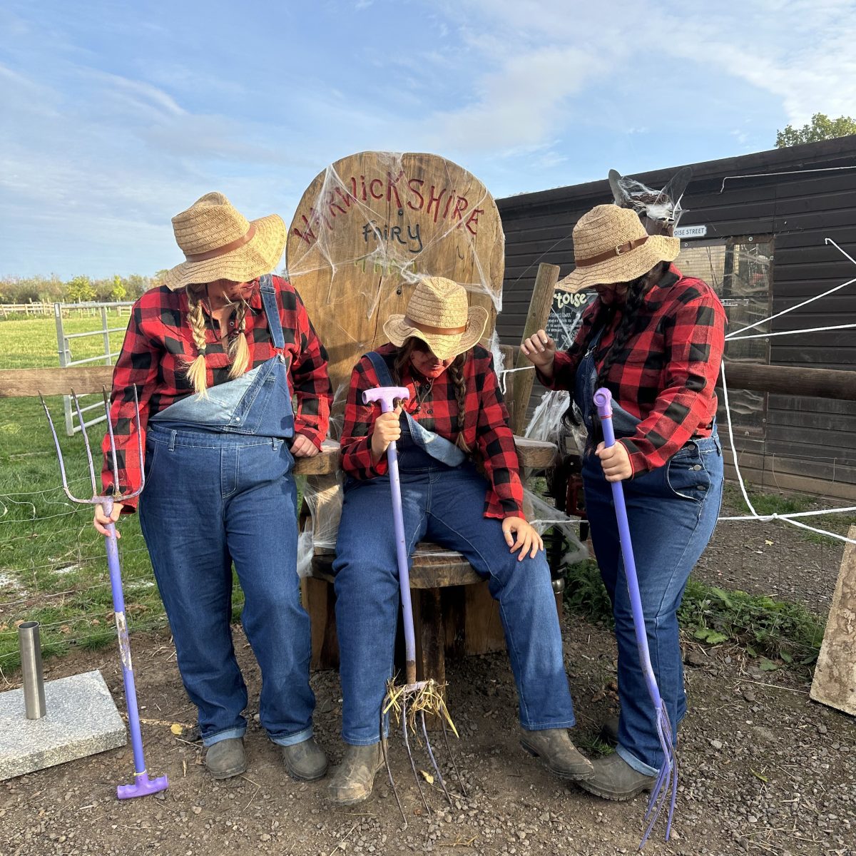 Three people in straw hats and plaid shirts posing with pitchforks in a rural setting.