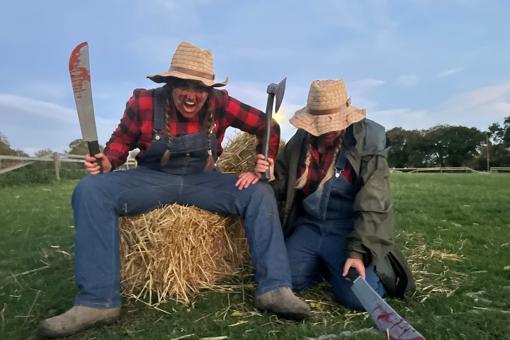 Two people in farm attire with hats, one holding a bloody machete, another holding an axe, sitting on hay.
