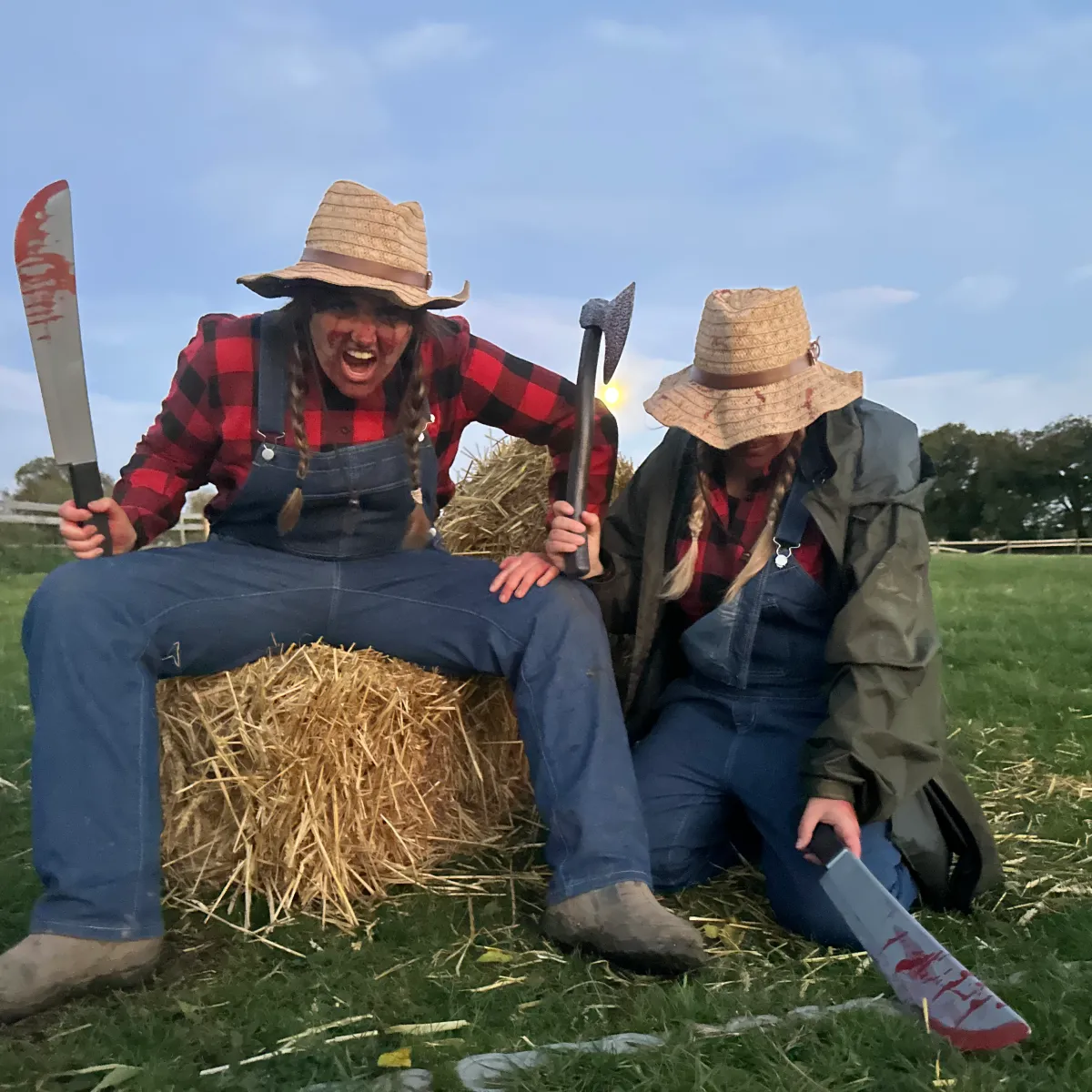 Two people in farm attire with hats, one holding a bloody machete, another holding an axe, sitting on hay.