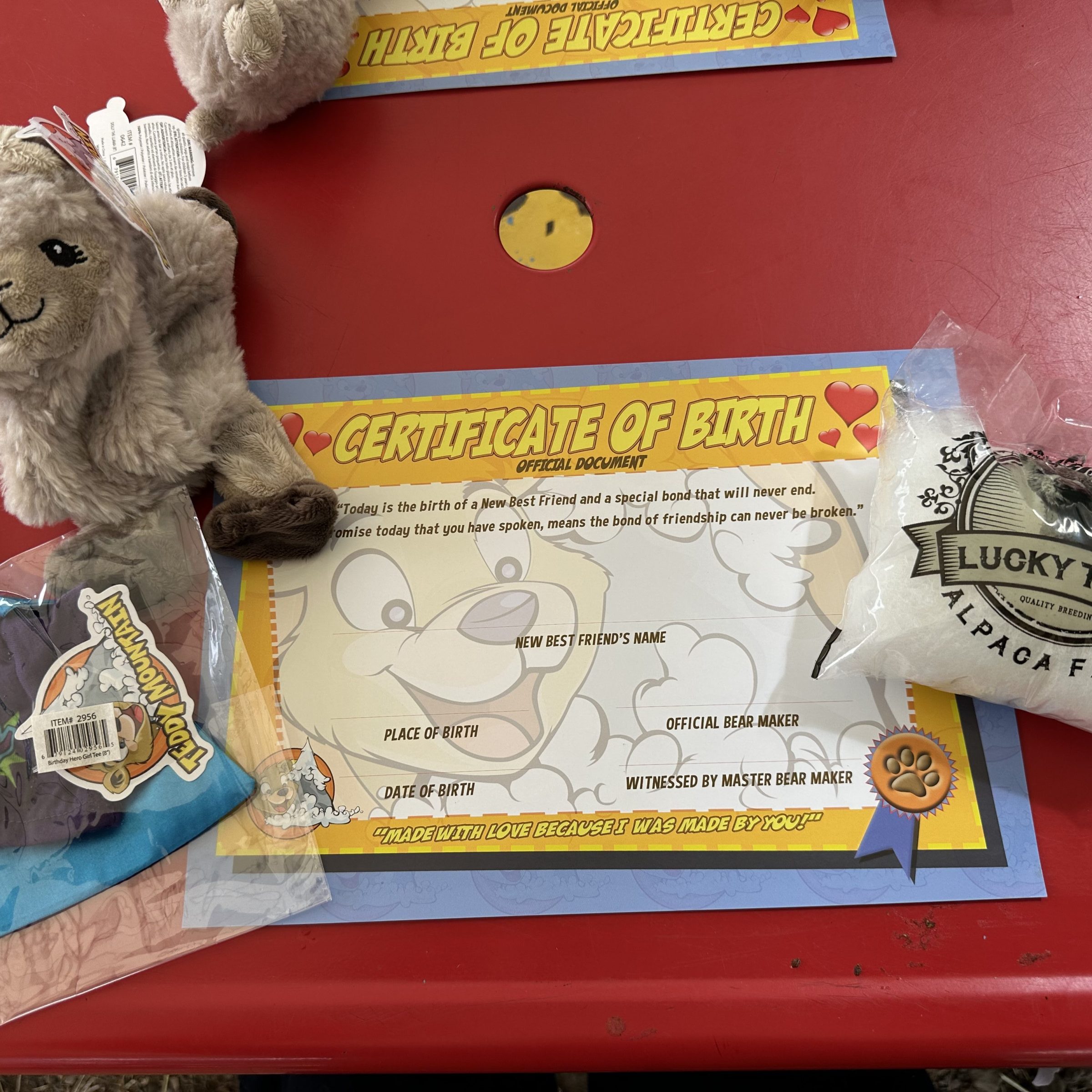 Stuffed alpaca toy with birth certificate and wool bag on red table.